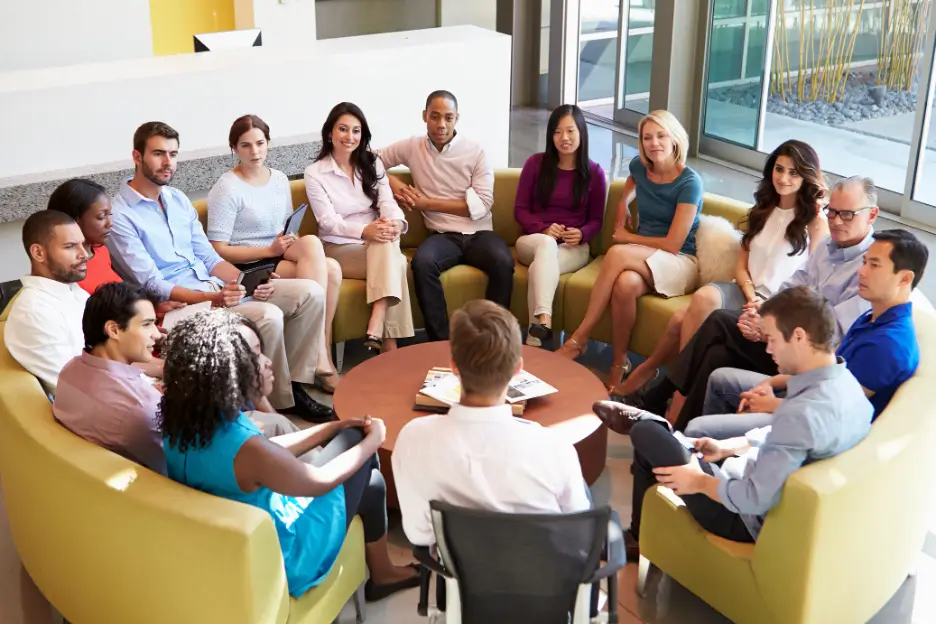 A diverse group of professionals sit in a circle in a modern office lounge, participating in a group discussion.